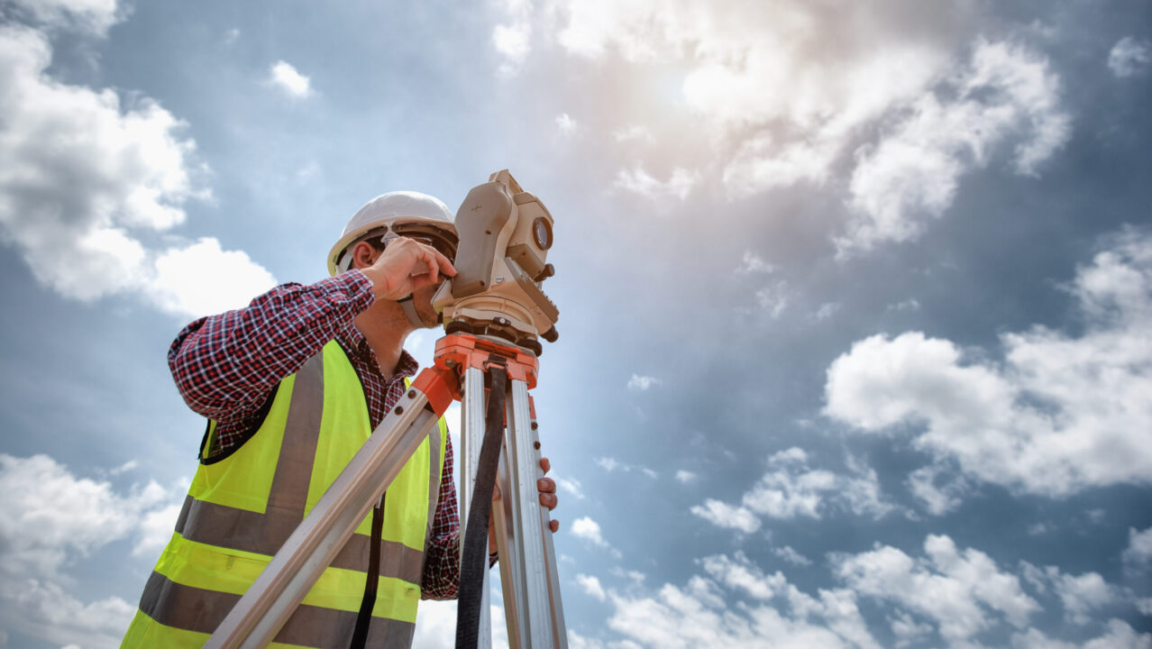 Persoon met witte veiligheidshelm en reflecterend vest bedient een landmeetkundig instrument op een statief. De persoon wijst omhoog terwijl hij door het apparaat kijkt. Op de achtergrond een blauwe lucht met wolken