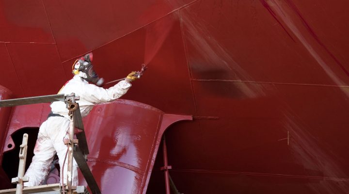 Werknemer in beschermpak brengt coating aan op scheepsromp, mogelijk blootgesteld aan Chroom VI-houdende stoffen