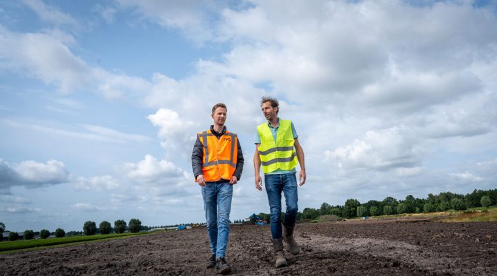 Twee personen in veiligheidsvesten lopen over een modderige bouwplaats in een landelijk gebied, met bomen en een deels bewolkte lucht op de achtergrond