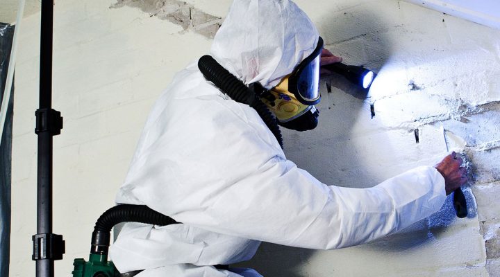 Inspecteur in beschermend pak en masker onderzoekt beschadigde muur met zaklamp tijdens asbestinventarisatie in een mogelijk risicovolle omgeving