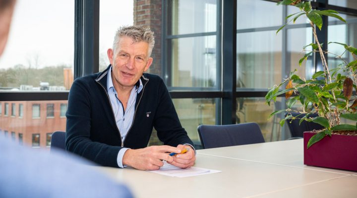 Brandveiligheidsadviseur in gesprek aan tafel, met documenten en uitzicht op gebouwen door groot raam