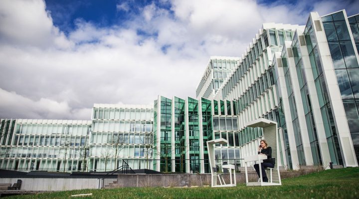 Modern glazen gebouw met op de voorgrond een persoon op een witte bank op een grasveld
