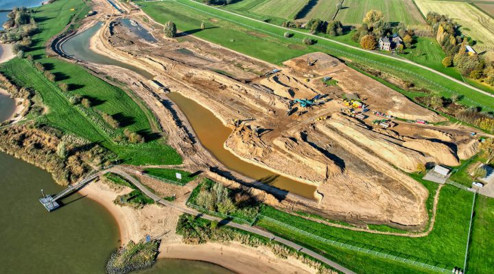 Luchtfoto van dijkverbeteringsproject met grondverzet en machines langs water, gericht op versterking en waterveiligheid