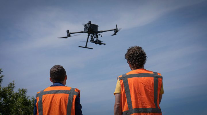 Drone Crew in veiligheidsvest observeert vliegende drone in buitengebied – inzet van drones voor inspectie van infrastructuur en moeilijk bereikbare locaties