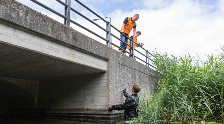 nspectieteam voert visuele controle uit bij brugconstructie met duiker in water en toezichthouders op brug – inspectie van civiele kunstwerken in natte omgeving