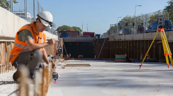 Landmeter gebruikt meetapparatuur op bouwplaats bij tunnelconstructie, met statief en apparatuur op de achtergrond