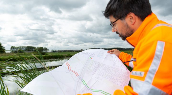 Medewerker in oranje veiligheidsjas bekijkt kaart in buitengebied met water en groen – planning en uitvoering van natuurontwikkelingsprojecten
