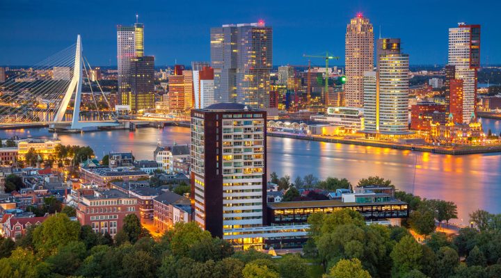 Nachtelijk stadsgezicht van Rotterdam met verlichte hoogbouw, Erasmusbrug en reflecties in de Nieuwe Maas; op de voorgrond lage bebouwing en groen