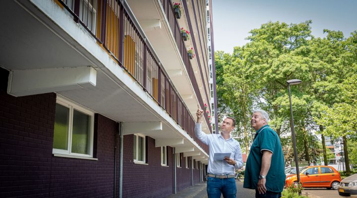 Twee personen inspecteren een woongebouw; één wijst omhoog, de ander houdt een clipboard vast. Op de achtergrond balkons, bomen en geparkeerde auto's